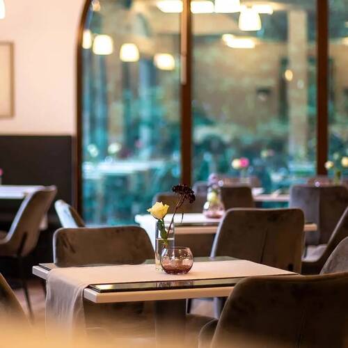 Stilvoll gedeckter Tisch im Bistro des Hotel Kastanienhof Erding mit Blumendekoration, gemütlicher Bestuhlung und Blick durch große Fensterfront.