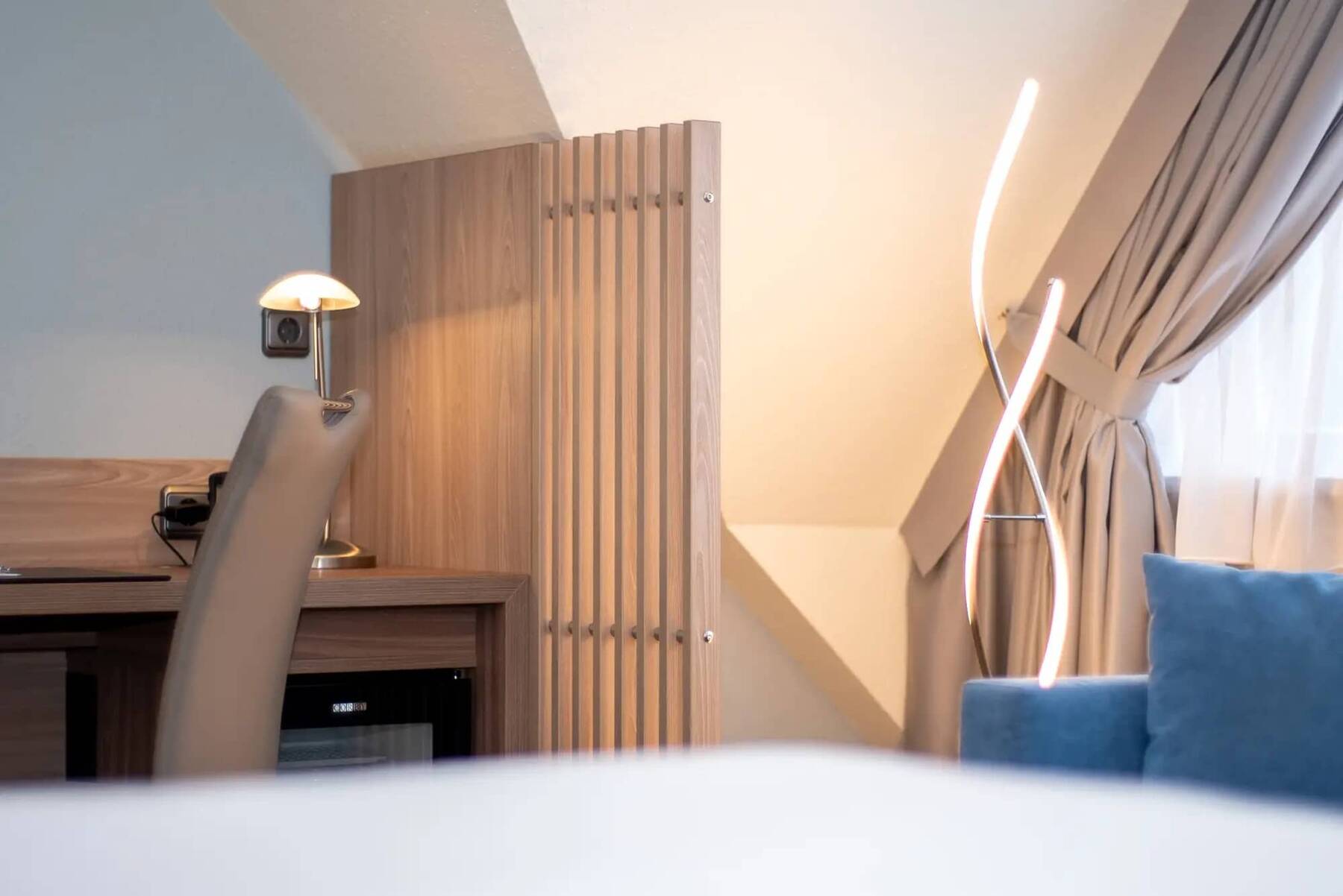 Helles Hotelzimmer im Kastanienhof Erding mit Schreibtisch, moderner Stehlampe, beigem Stuhl und gemütlicher Sitzecke am Fenster.
