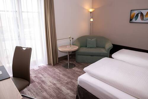Double room in the family suite at the Hotel Kastanienhof Erding with armchair, desk, cosy double bed and natural light through floor-to-ceiling windows.