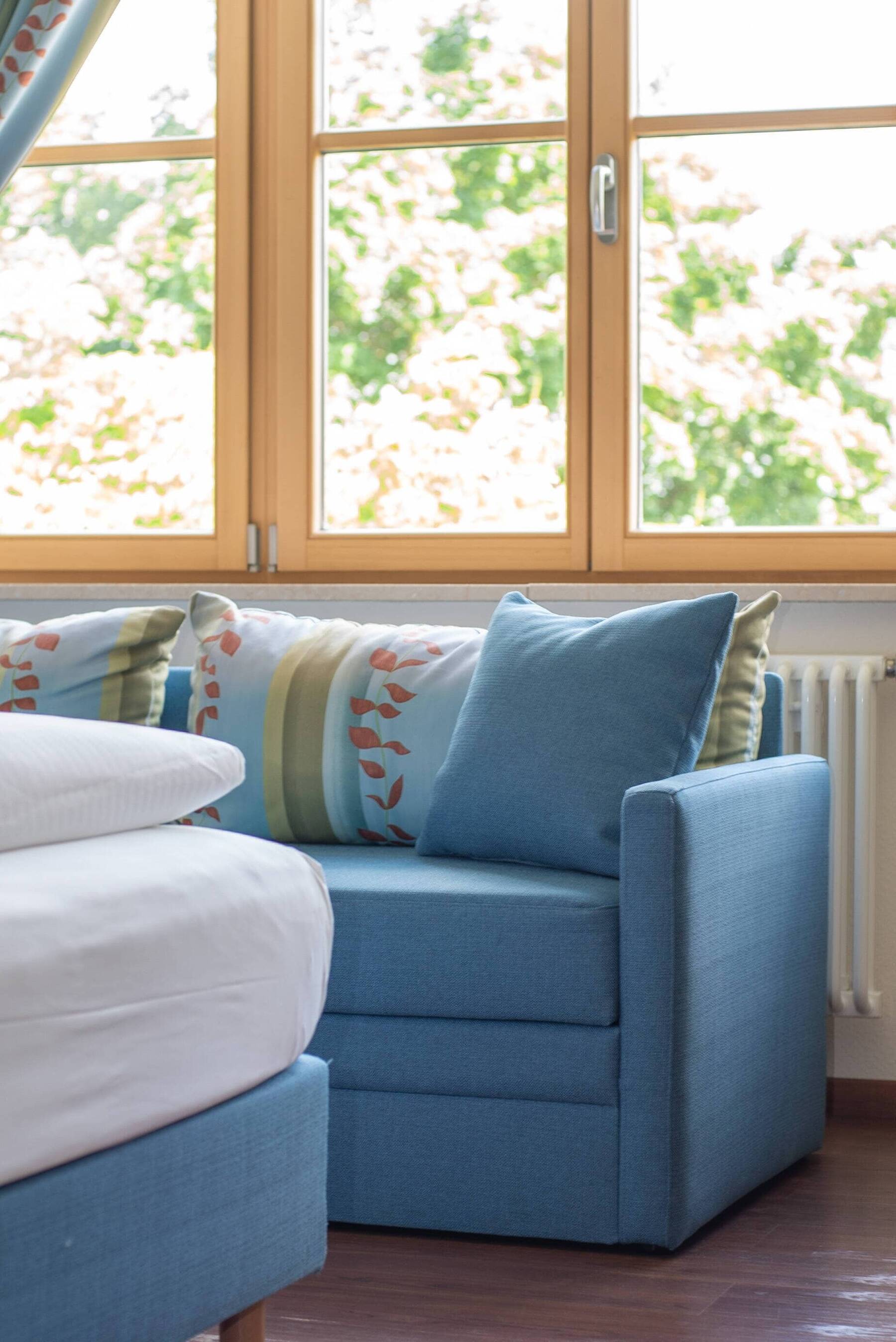 Gemütliche, pastell-blaue Couch mit bunten Kissen und hellem Fenster dahinter im Doppelzimmer Executive.