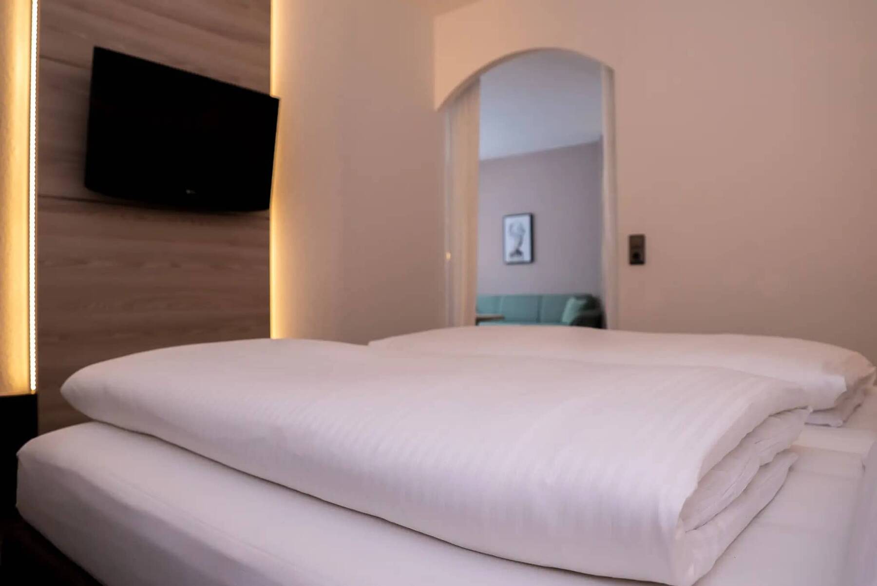 Modernes Schlafzimmer mit großem Bett und einem Flachbildfernseher an einer Wand mit Holzpaneele und warmen Lichtstreifen dahinter sowie Blick in das angrenzende Wohnzimmer mit Sofa.