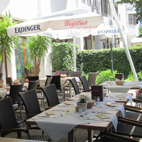 Sonnige Terrasse am Hotel Kastanienhof Erding mit gedeckten Tischen, gemütlichen Rattanstühlen und Erdinger Sonnenschirmen in grüner Hoflage.