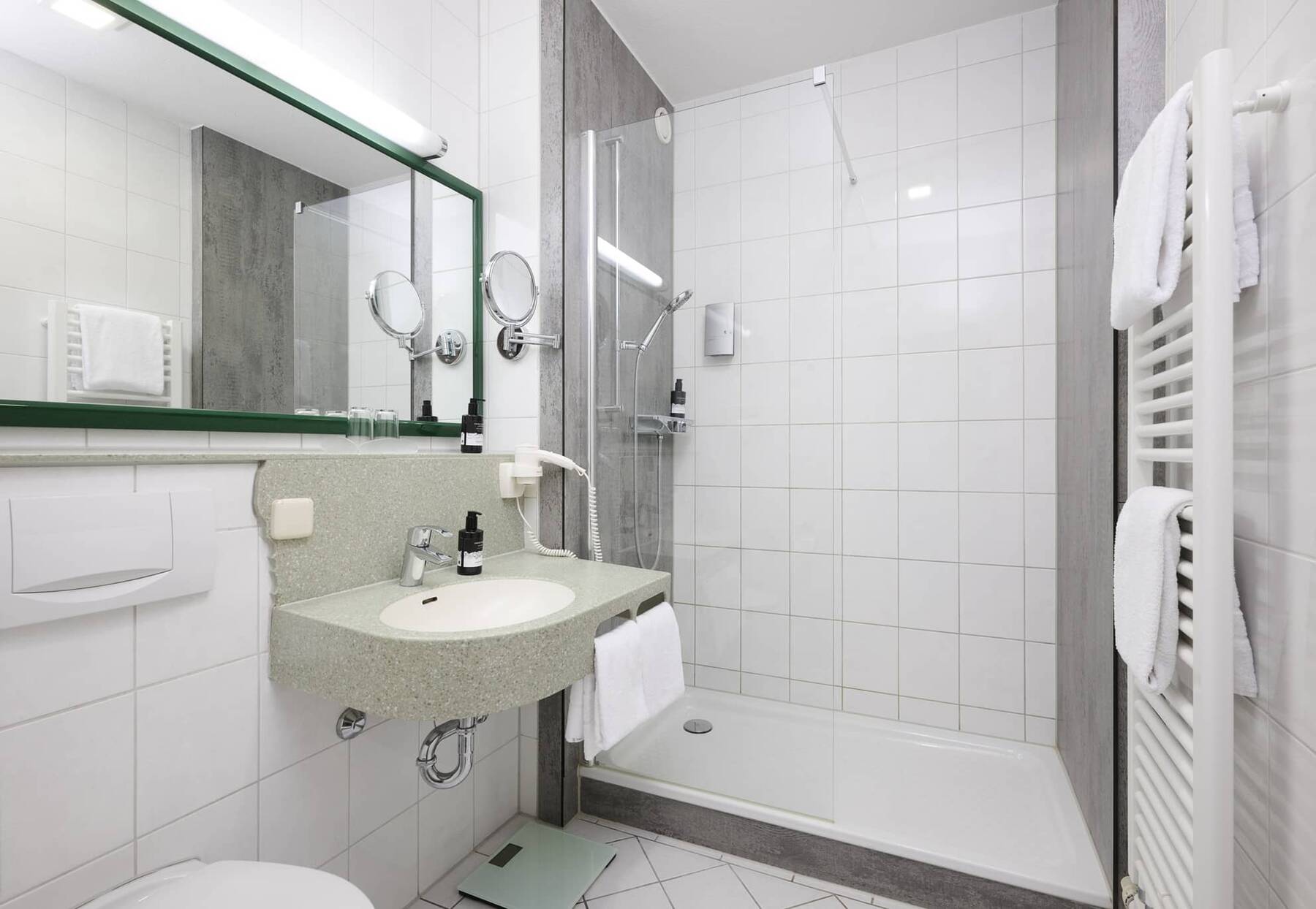 Helles Badezimmer des Superior Doppelzimmers im Hotel Kastanienhof Erding mit Dusche, Waschbecken, großem Spiegel und Handtuchheizkörper.