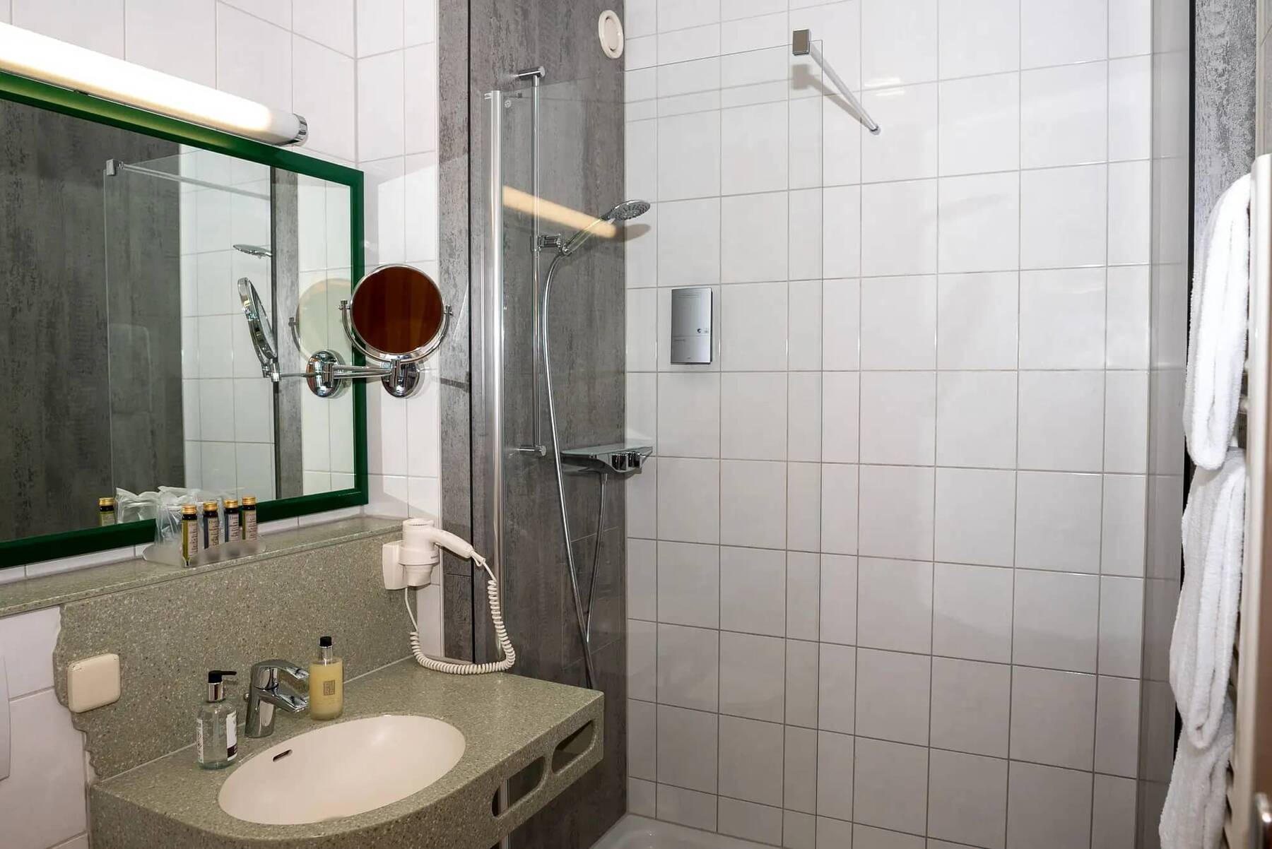 Das Bad im Doppelzimmer des Hotels Kastanienhof mit Dusche, einem grün designten Waschbecken, Spiegel, Schminkspiegel, Föhn und Kosmetikartikel.