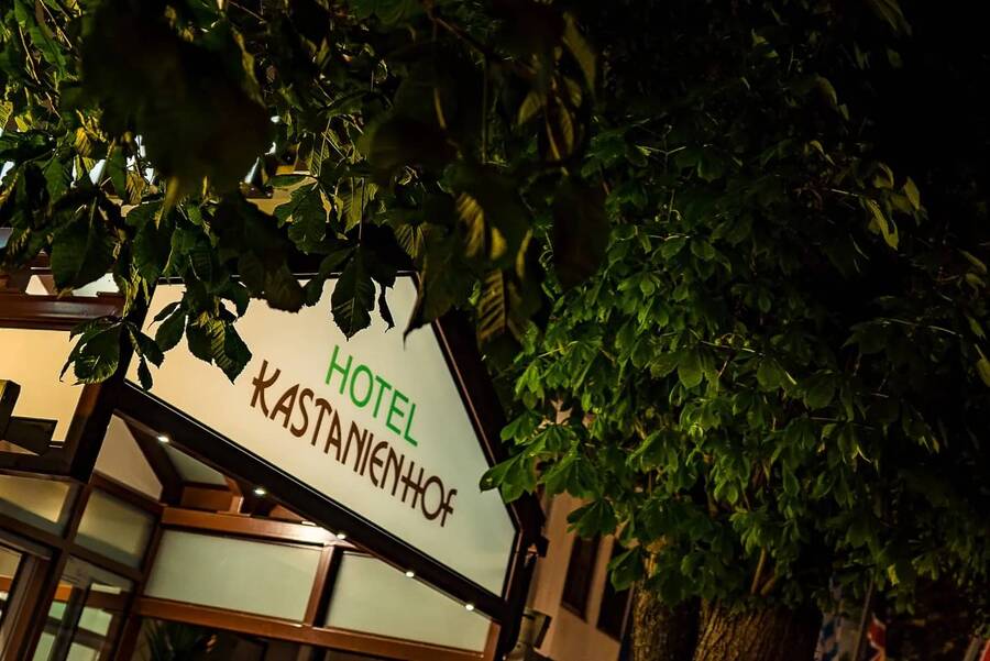 Nächtliche Außenaufnahme des Eingangs vom Hotel Kastanienhof in Erding, eingerahmt von dichtem Kastanienlaub.