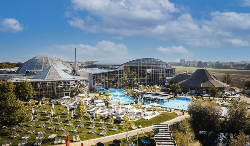 Luftaufnahme der Therme Erding – ideales Ausflugsziel für Gäste des Hotels Kastanienhof: Weitläufiges Freizeitbad mit mehreren großen Glaskuppeln und Gewächshausstrukturen, türkisfarbenem Außenpool mit Liegestühlen und weißen Sonnenschirmen auf gepflegter Grünfläche – perfekt für einen Kurzurlaub nahe Erding, umgeben von Feldern und bewölktem blauem Himmel.
