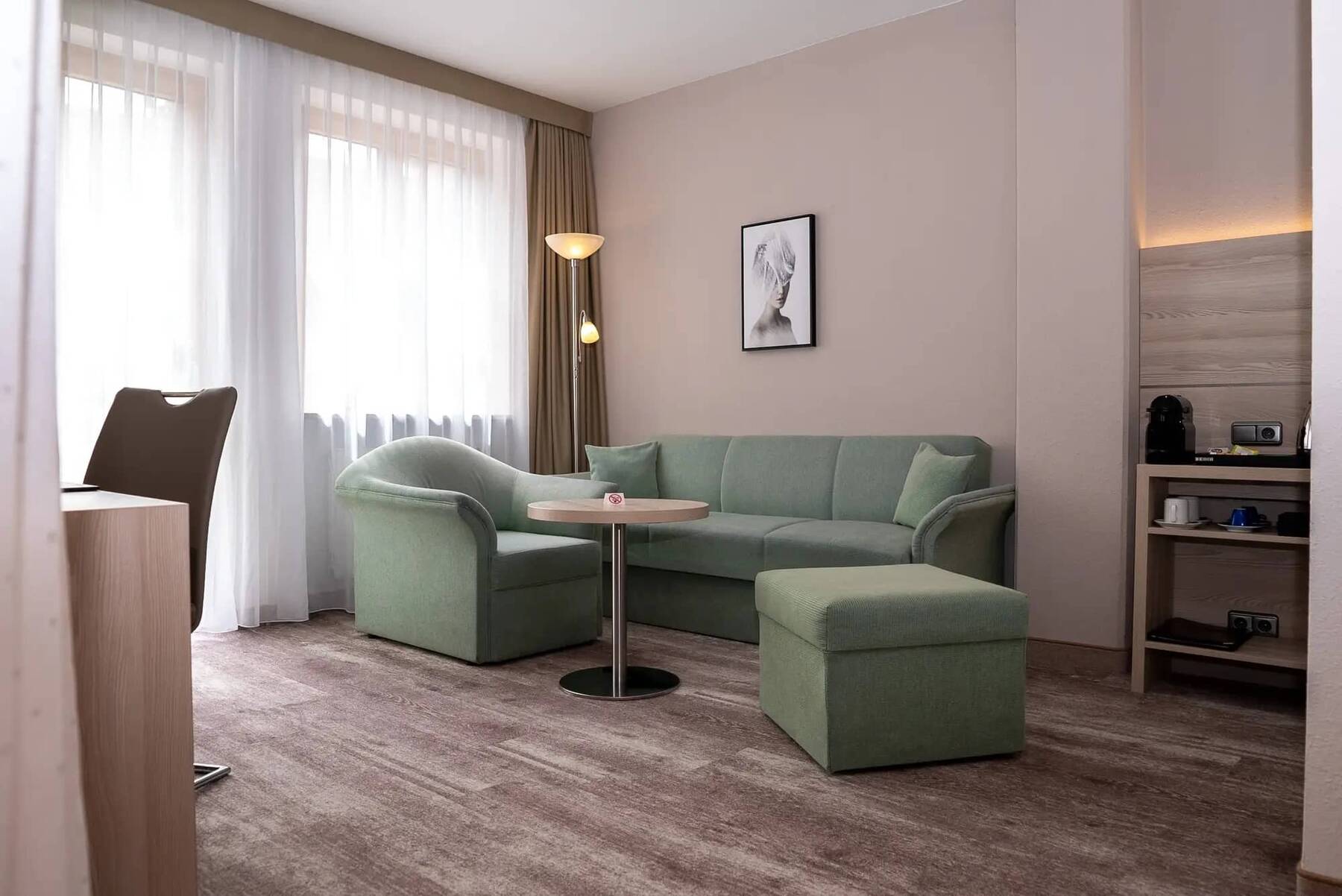 Geräumige Sitzlounge mit pastellgrüner Couch und zwei Sesseln, bodentiefen Fenstern und rechts daneben eine Tee-Ecke.