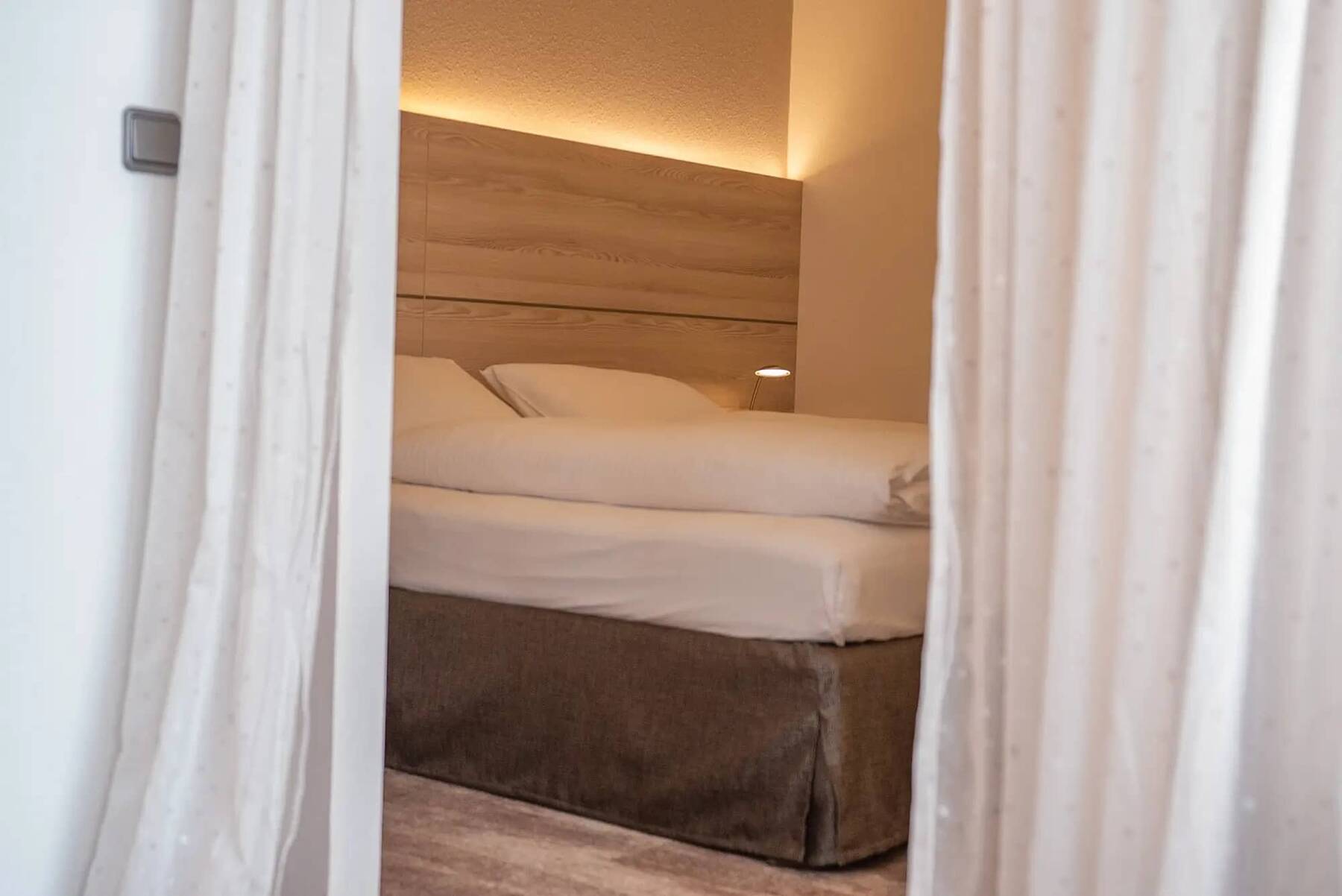 Einblick in das Schlafzimmer der Junior Suite im Hotel Kastanienhof Erding mit Doppelbett, Holz-Kopfteil, indirekter Beleuchtung und Vorhang als Raumtrenner.